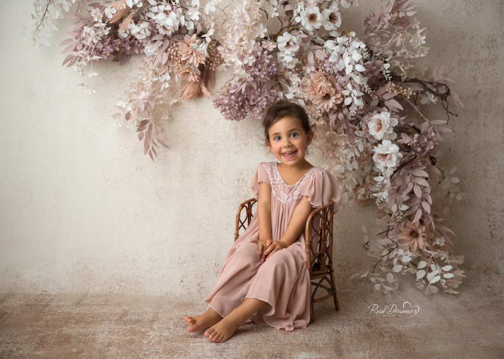 una bambina su seggiolina di legno e sfondo con fiori durante il servizio fotografico bambini