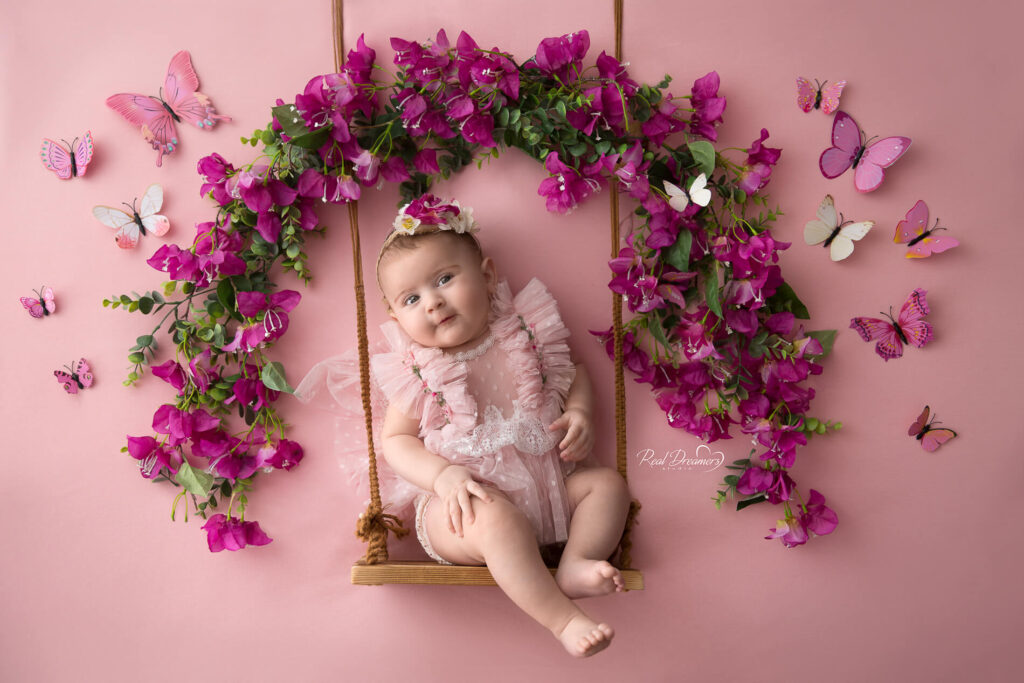un bebe su un'altalena con intorno fiori e farfalle durante un servizio fotografico bebe