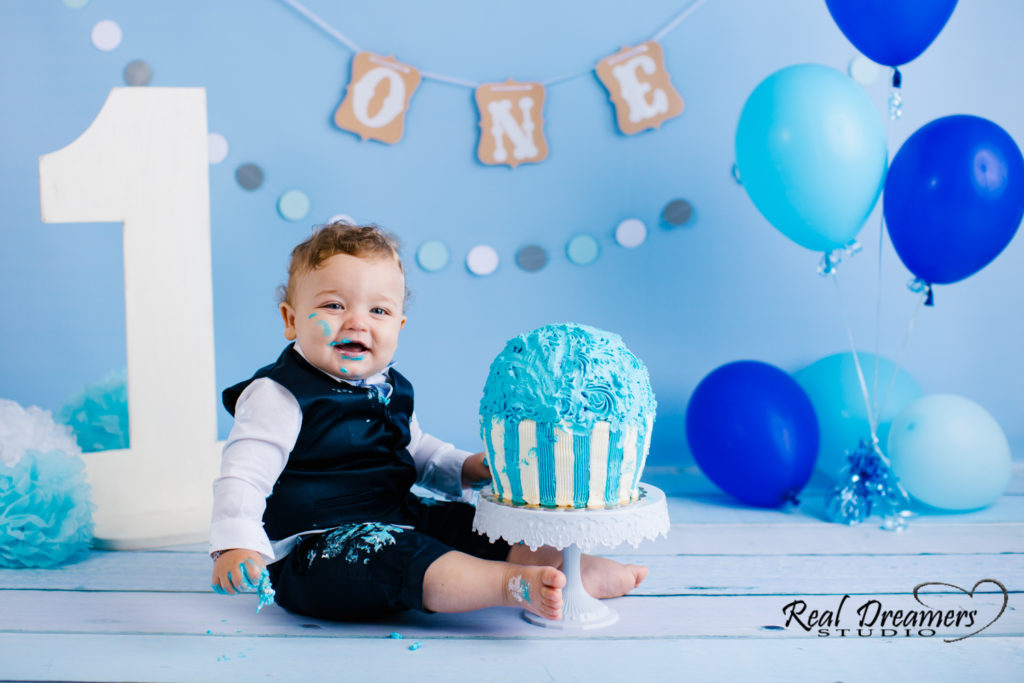 un bimbo con fondale di palloncini e numero uno, torta davanti e bocca sporca di panna durante il suo servizio fotografico smash cake